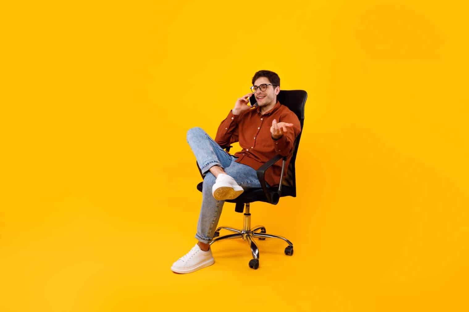 Business Man Talking On Phone Sitting In Chair Over Yellow Studio Background. Man Communicating Having Pleasant Cellphone Conversation. Modern Mobile Communication Concept. Full Length Shot