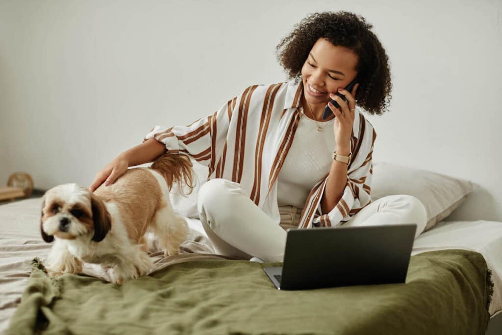 Unverblümtes Porträt einer schwarzen jungen Frau, die von zu Hause aus auf dem Bett arbeitet und mit ihrem Hund spielt, Kopierraum - psychische gesundheit