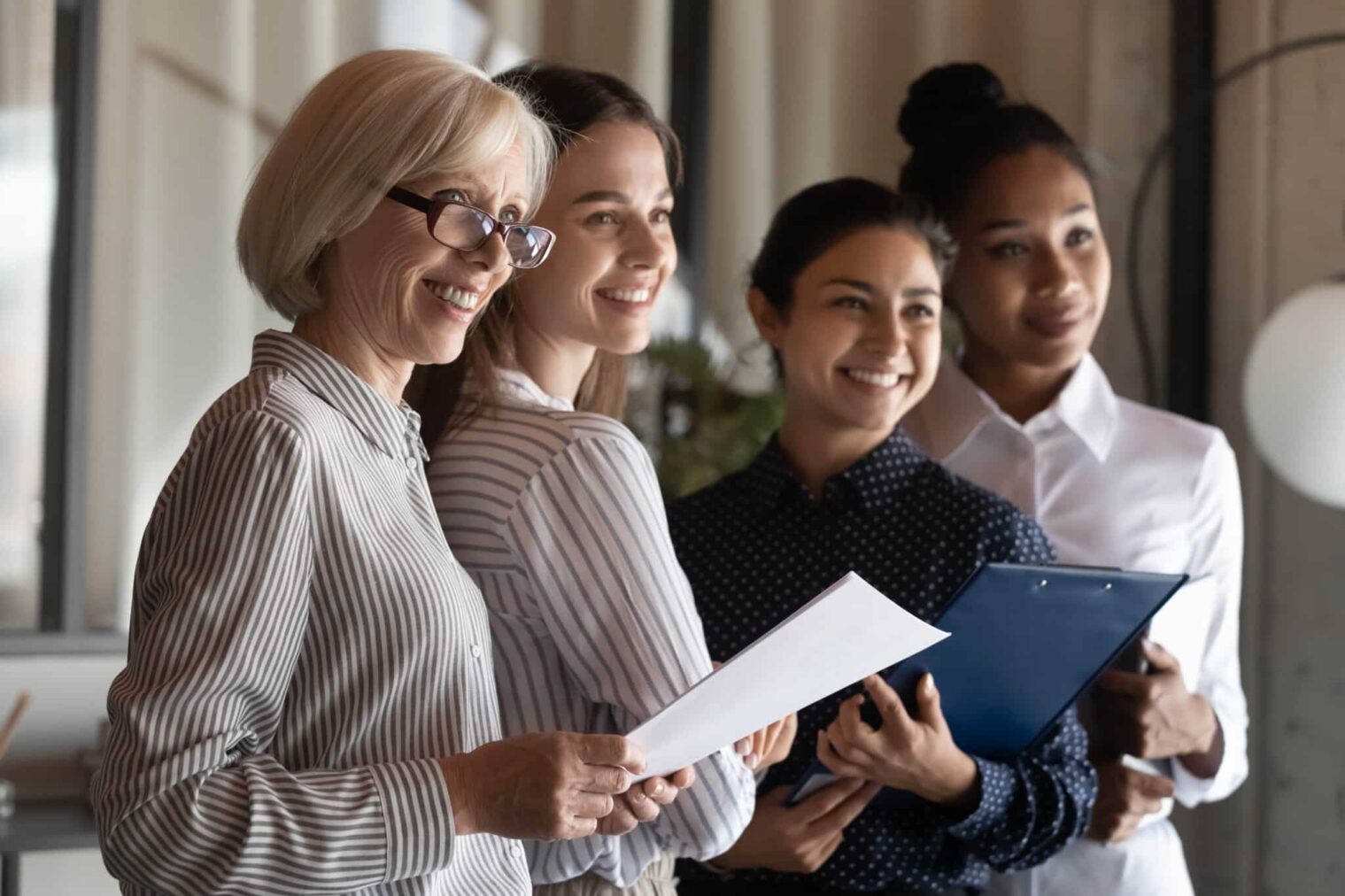 Motivierte multirassische weibliche Team-Büroangestellte verschiedener Generationen schauen auf eine Seite und lächeln, bereit, Kunden zu helfen. Professionelle verschiedene Geschäftsfrauen stehen zusammen halten Dokumente Klemmbretter