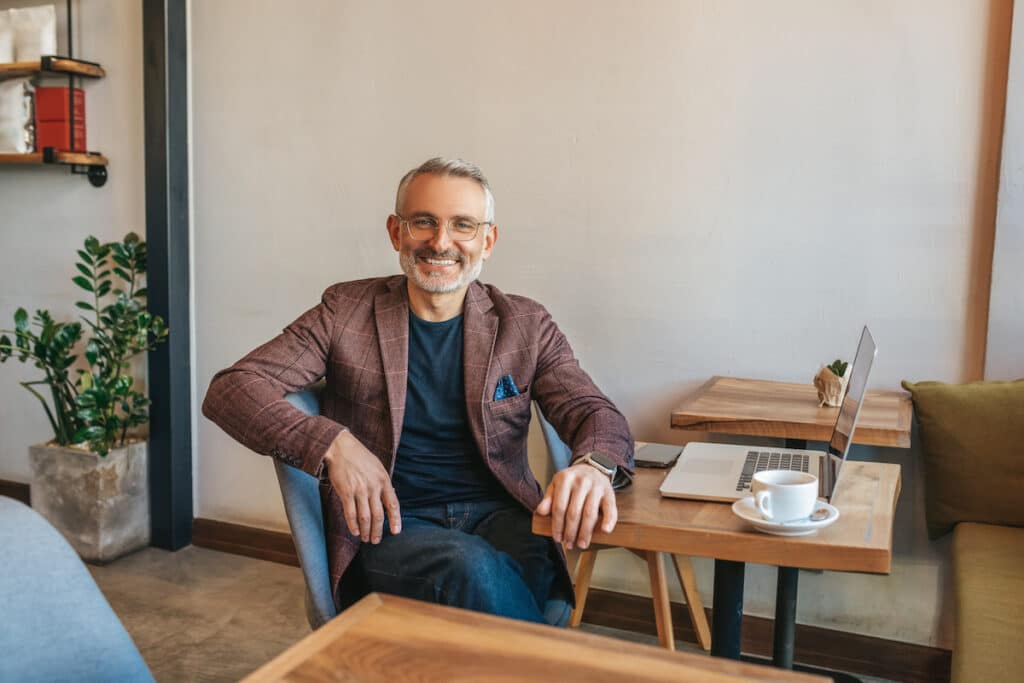 Grauhaariger bärtiger Mann mit Brille sitzt am Tisch mit Laptop und Kaffee und lächelt in die Kamera drinnen am Tag - Generation