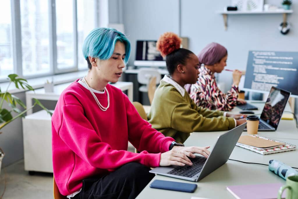 Ein buntes Team von Softwareentwicklern der Generation Z arbeitet in einem Büro und konzentriert sich auf einen asiatischen jungen Mann mit gefärbten Haaren, der einen Computer im Vordergrund benutzt.