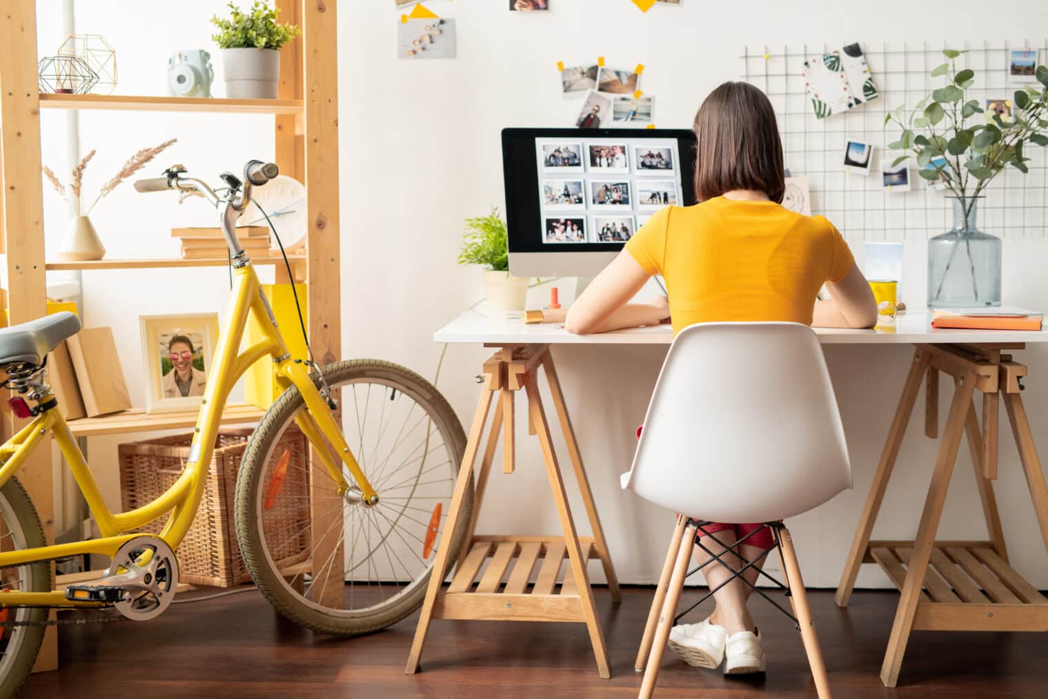 Rear view of a young brunette woman sitting on a white chair in front of a computer monitor, browsing through a collection of photos -