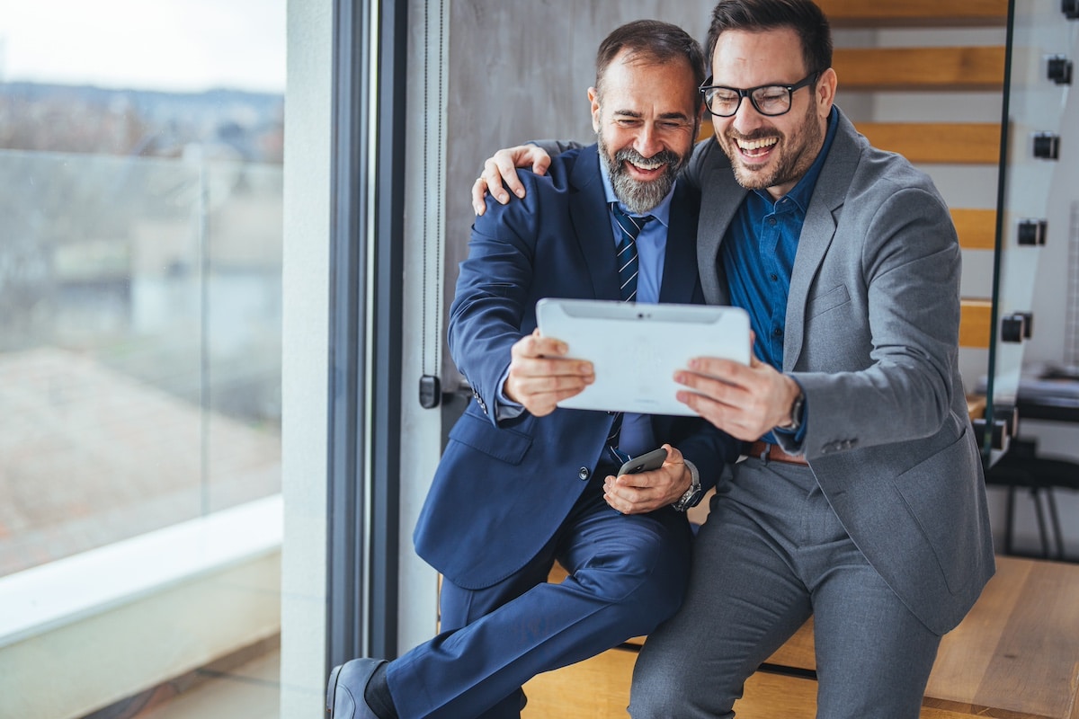 Fröhlicher und lächelnder junger erfolgreicher männlicher Geschäftsmann steht mit einem Kollegen vor einem Tablet-PC in einem modernen Büro und Coworking Space. Kollegen verwenden Tablet-PC -