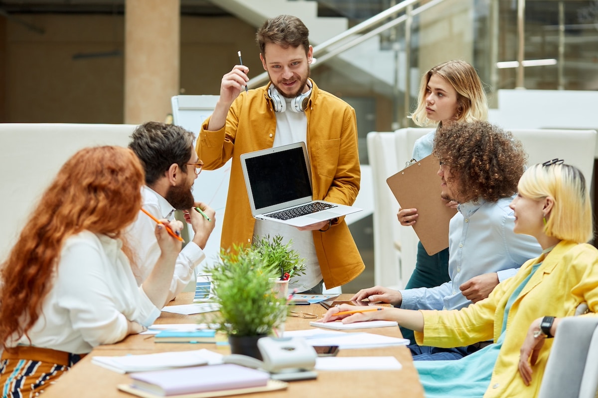 Fröhliche positive Gruppe von talentierten Arbeitern hört einem bärtigen jungen Mann mit Notizbuch zu, drückt Aufmerksamkeit aus, Startup-Konzept -