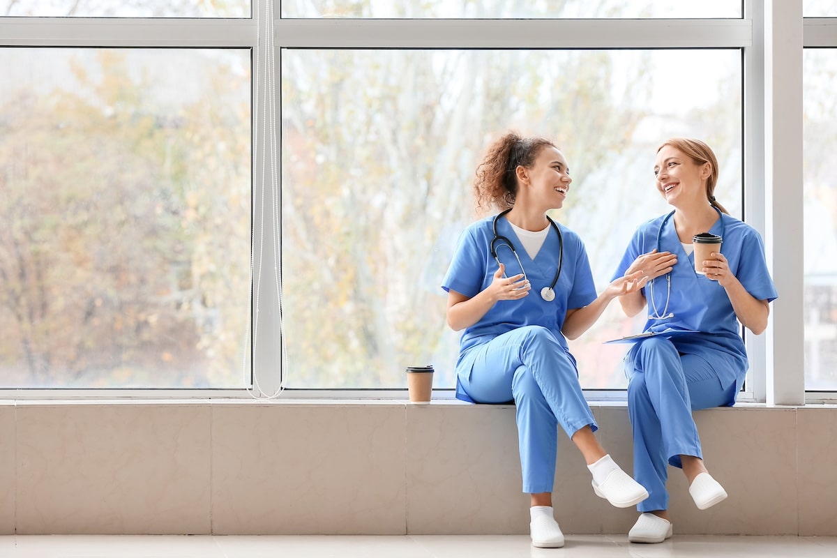 Female doctors drinking coffee while sitting on window sill in clinic - Empowering caregivers