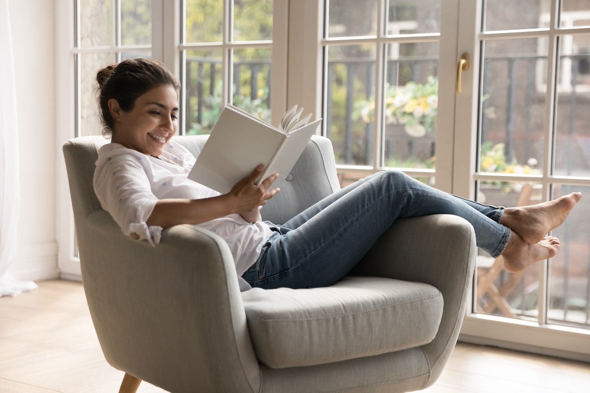 Young 25s smiling Indian woman reading novel book relaxing alone on comfort armchair in cozy room, spend leisure enjoy favourite literature bought in bookshop. Booklover pastime at home, hobby concept -