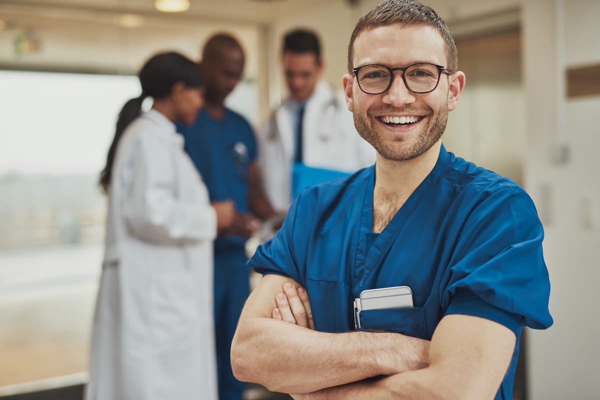 Smiling optimistic young male hospital surgeon standing in front of his colleagues with folded arms beaming at the camera - reduce stress