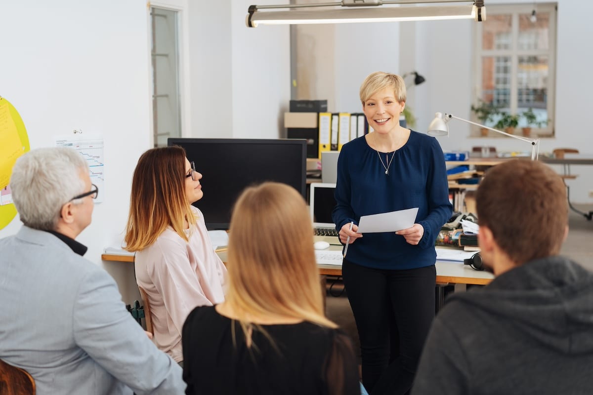 Woman addressing her speech to a group of her colleagues at business meeting, standing in front of them with a sheet of paper and smiling while talking