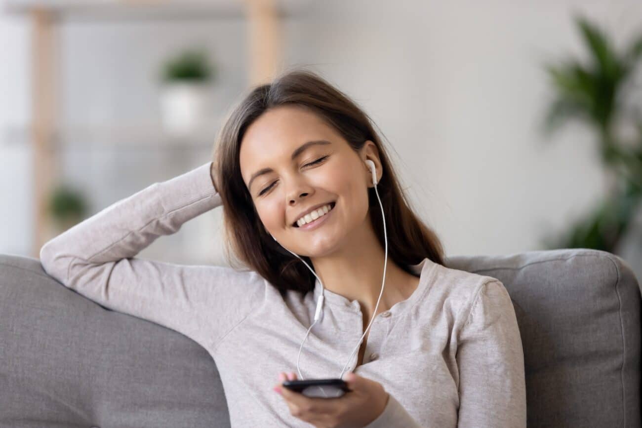 Attractive woman in casual clothes wearing earphones sitting on couch holding phone closed eyes smile enjoy popular tracks songs resting at home feels good. Leisure activities favourite music concept -