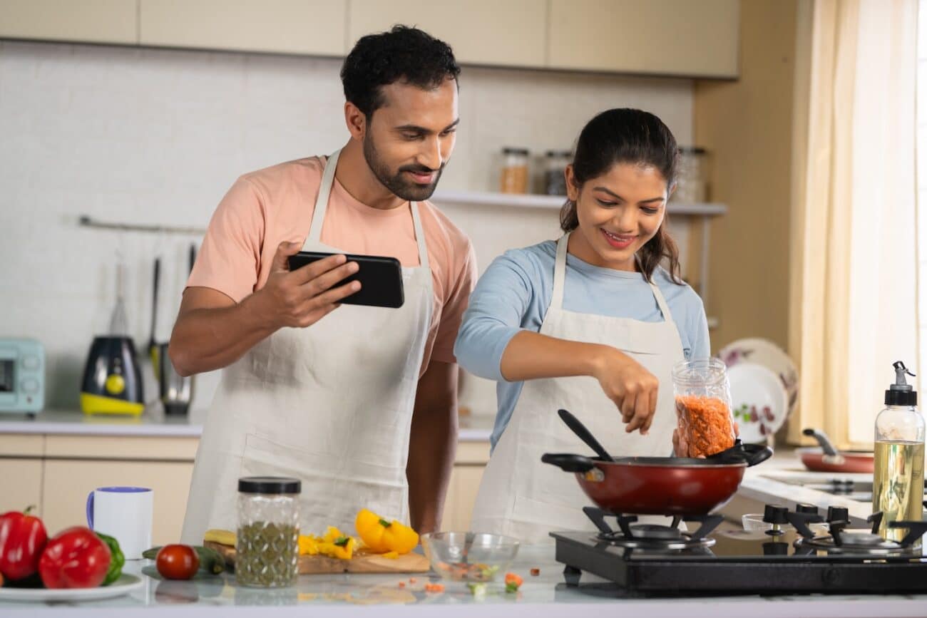 Joyful Indian couples cook or trying new food recipe by watching online videos from Mobile phone at Kitchen - concept of technology, relationship bonding and collaboration. -