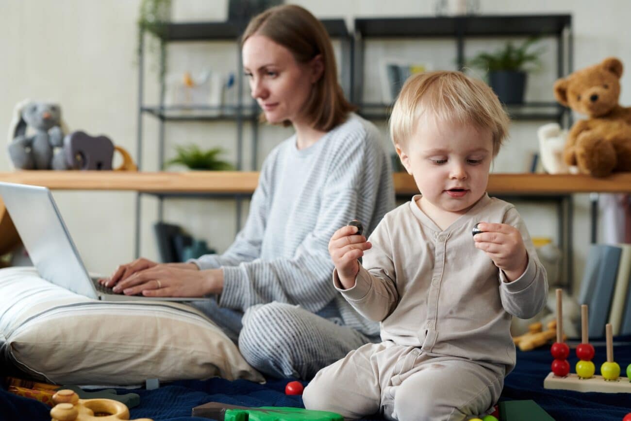 Cute little child playing toys on bed with his working mother using laptop for online work in background - workplace health management