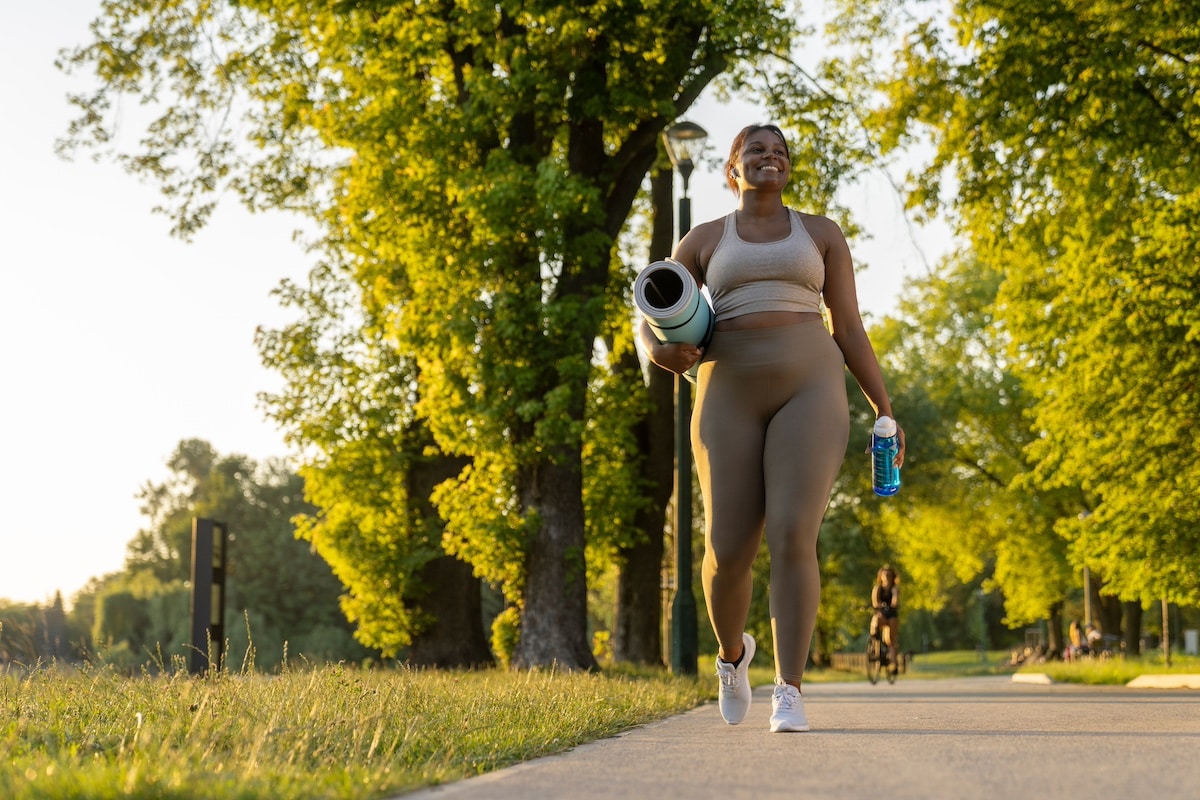 Young African American woman walking with exercise mat and bottle of water through the park in a summer day -