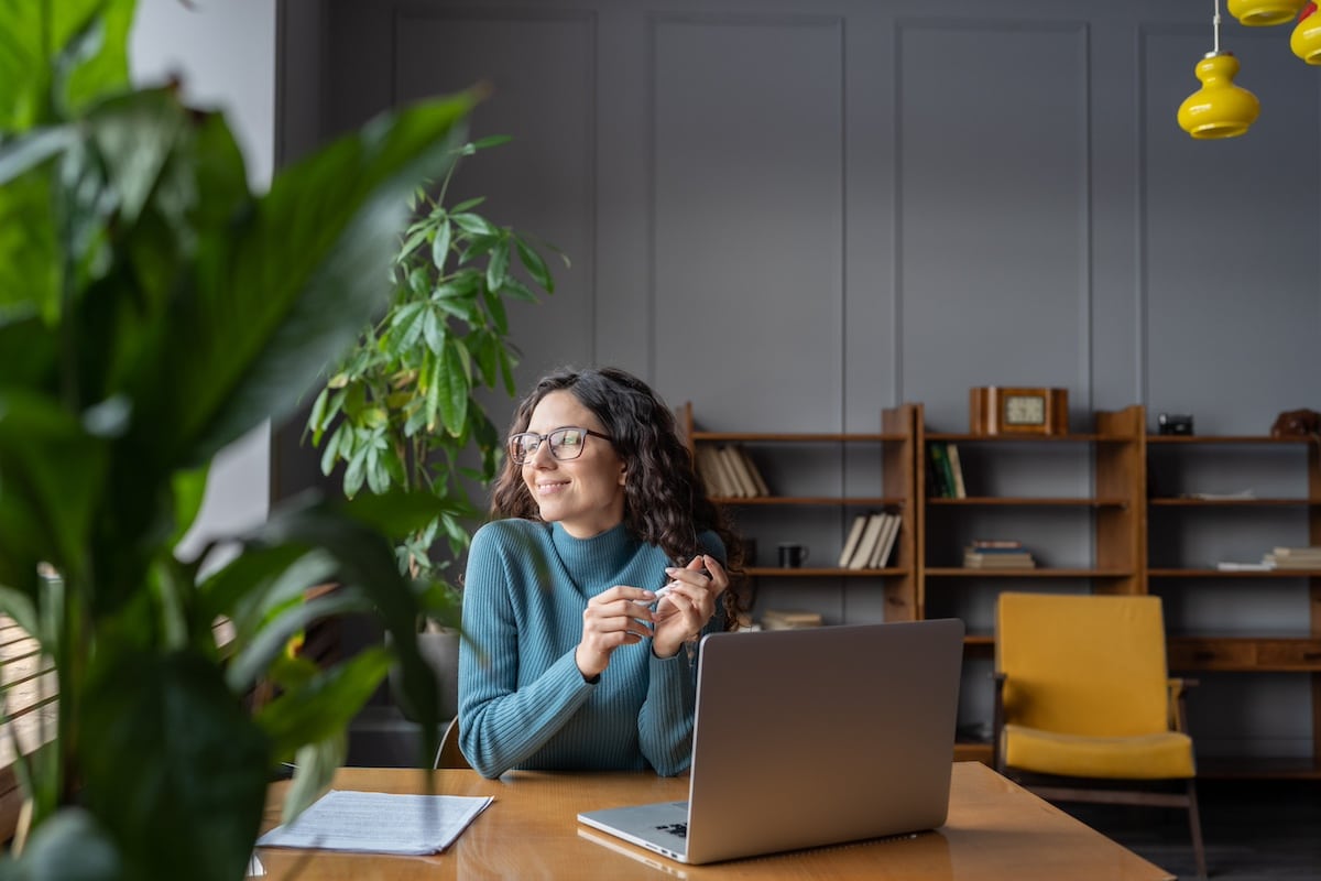 Happy female employee looking in window with satisfied face expression while sitting in office in front of laptop, woman taking break from computer work at workplace. Job satisfaction concept