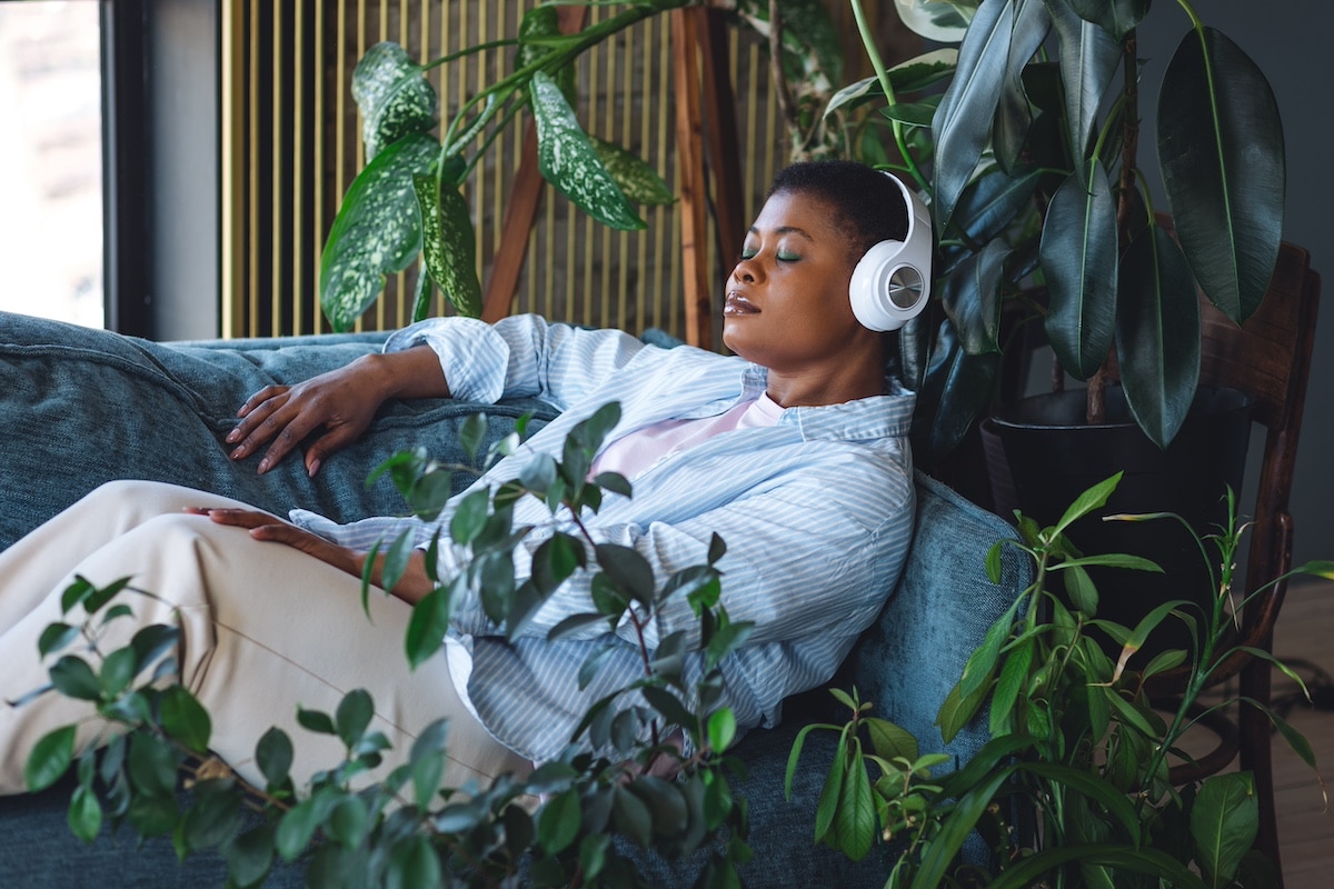 Concept of relaxation and meditation at home. Young plus size calm African American woman relaxing on the couch, listening to favourite music in headphones. Mindfulness, wellbeing, work life balance -