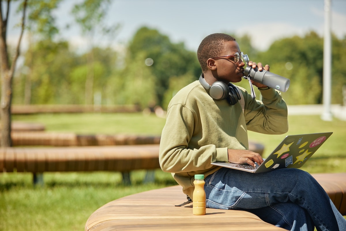 Side view of young African American male student on bench studying using laptop and drinking water to stay hydrated on sunny day, casual outdoor learning scene, copy space -