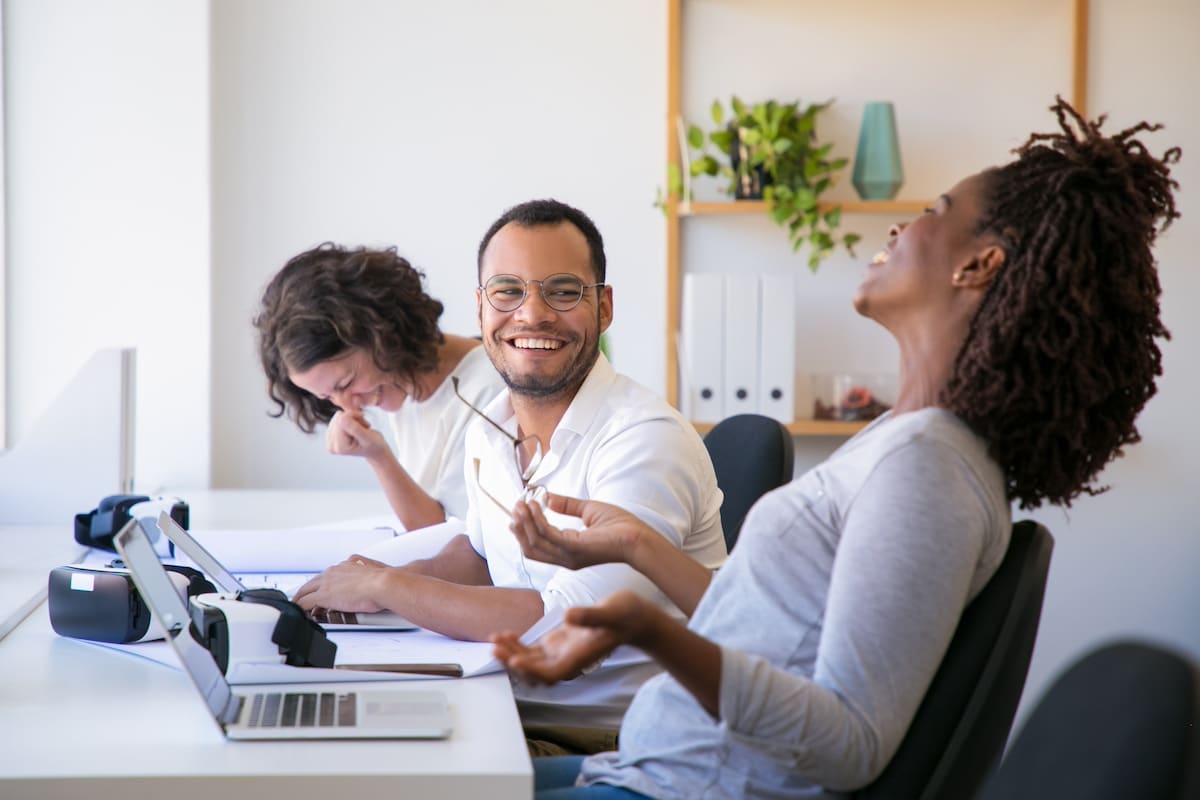 Diverse team of VR developers chatting while testing product. Man and women in casual sitting at table with virtual reality glasses and laptops, talking and laughing. Positive workgroup concept -