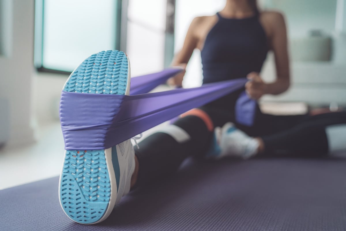 Resistance band exercise at home. Woman doing pilates workout using elastic strap pulling with arms for shoulder training on yoga mat indoors.