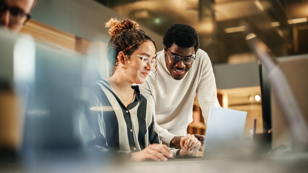 Portrait of Two Creative Colleagues Using Laptop to Discuss Work Project at Office. Young Black Technical Support Specialist Helping Female Customer Relationship Coordinator. Teamwork Concept