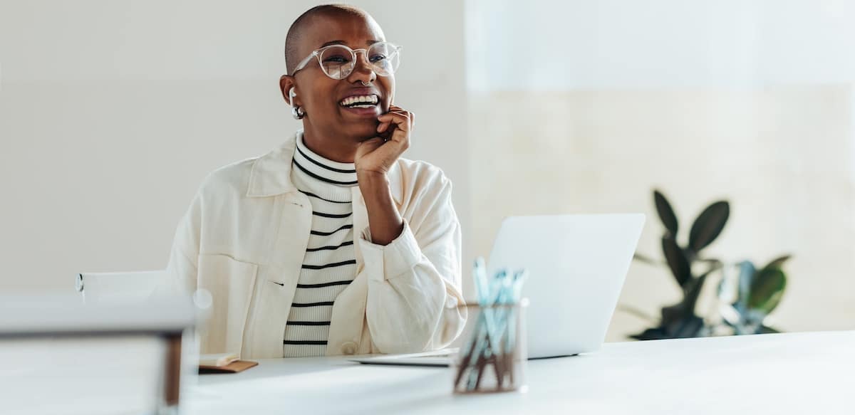 Eine junge, glückliche Geschäftsfrau feiert einen erfolgreichen Moment, während sie in einem hellen, modernen Büro an ihrem Laptop arbeitet und so positive Stimmung und Erfolg am Arbeitsplatz demonstriert. -