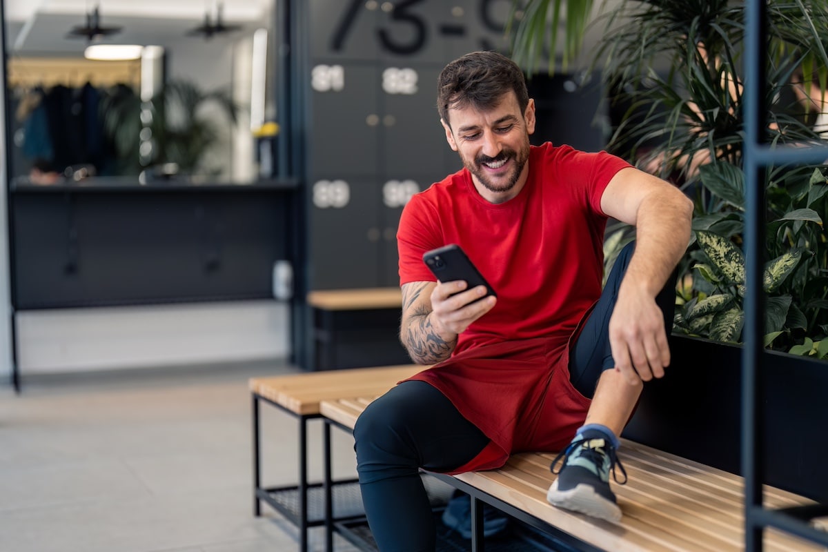 Handsome positive young sports man gym coach or client using smart phone technology fitness apps after workout tracking body wellness progress healthy lifestyle improvement while sitting in locker room. - employee wellbeing