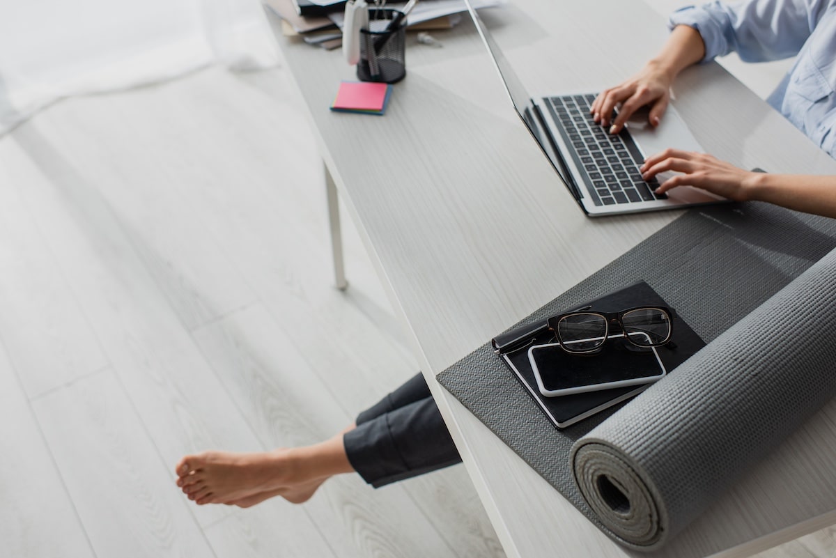 cropped view of barefoot businesswoman working on laptop at workplace with yoga mat, notepad, smartphone and eyeglasses - corporate health initiatives