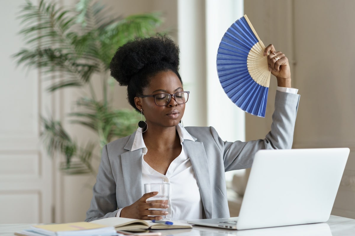 Eine erschöpfte Geschäftsfrau, die vor Hitze arbeitet und mit einem Papierfächer nach frischer Luft fächert. Eine afroamerikanische Geschäftsfrau aus der Generation der Millennials leidet unter der Hitze in Innenräumen im Büro oder zu Hause bei der Arbeit am Laptop. - Sommerblues am Arbeitsplatz