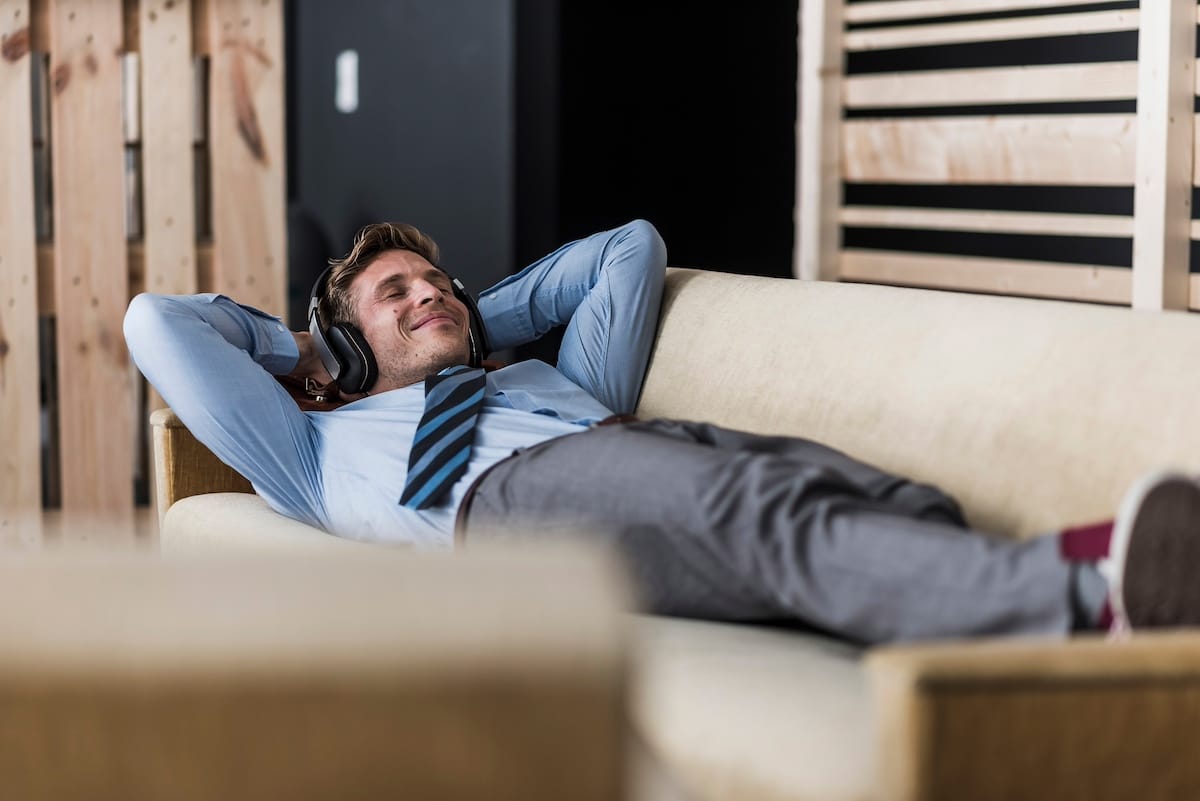 Businessman with headphones lying on couch in office lounge - mindfulness