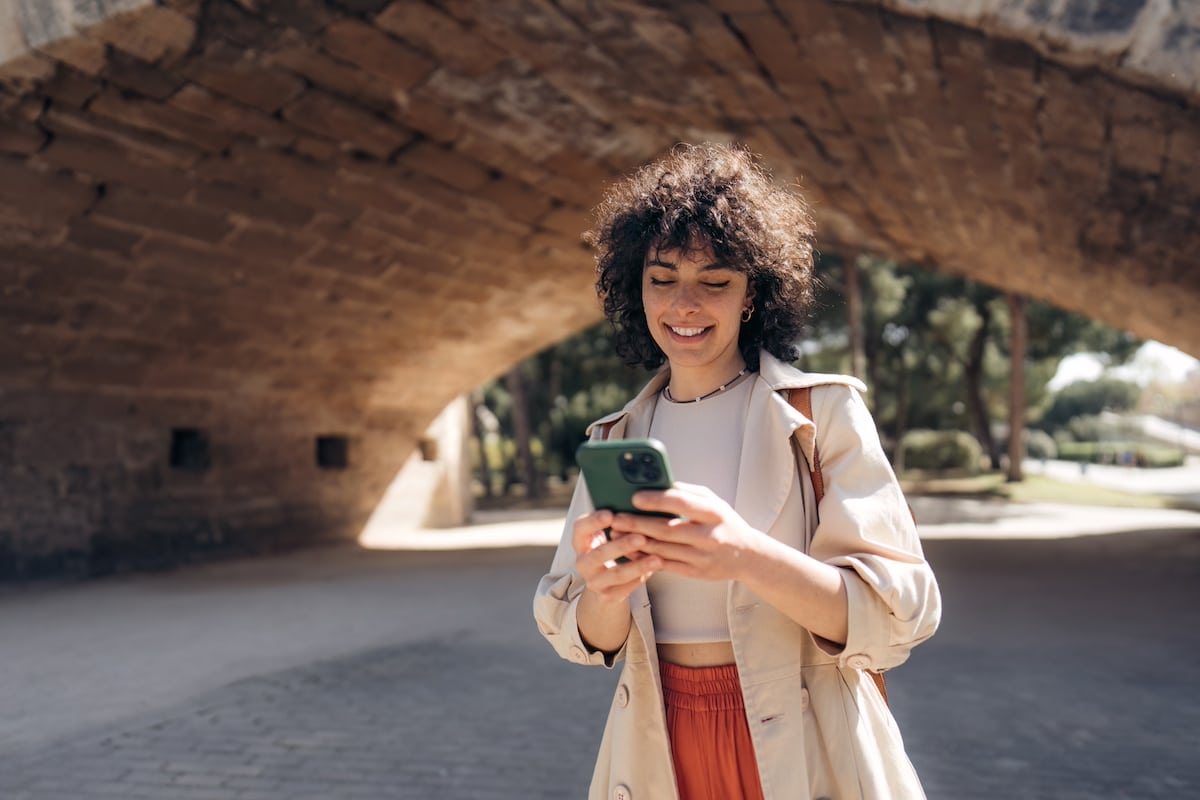 Young smiling woman wearing casual clothes holding smartphone using cellphone modern technology, looking at mobile, checking cell phone apps, texting, browsing internet while walking down the street. -