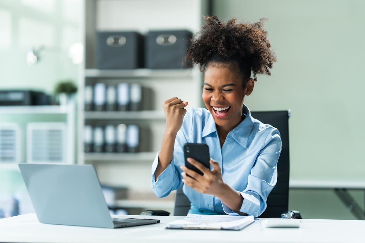 Exuberant and triumphant, a young African American woman in a blue formal shirt with afro brown hair operates a tablet, notebook, and mobile phone in a modern office setting. - corporate health initiatives