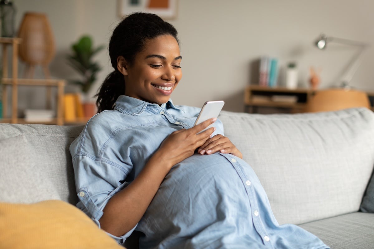 Happy Pregnant African American Woman Using Mobile Phone Texting Networking In Social Media Sitting On Couch At Home. Pregnancy Lifestyle, Gadgets And Communication Concept -