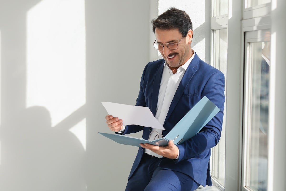 Ein fröhlicher erwachsener kaukasischer Geschäftsmann in einem blauen Anzug liest in einem hellen Büro Dokumente durch und drückt seine Zufriedenheit und Zuversicht in der Nähe eines Fensters aus, durch das Sonnenlicht hereinströmt. -