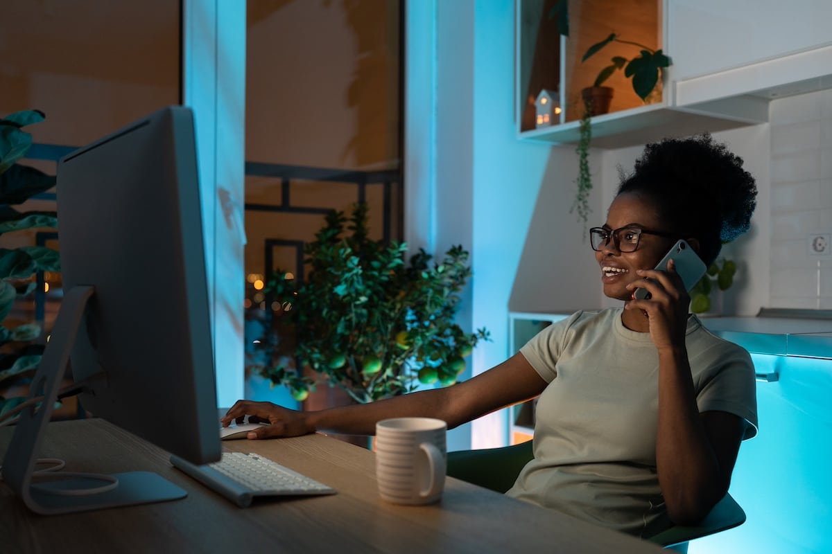 Positive, gemischtrassige Frau mit Brille, die eine Computermaus benutzt, mit dem Smartphone telefoniert und spät nachts im Homeoffice an einem Desktop-PC arbeitet. Konzept der Fernarbeit - Sommerblues am Arbeitsplatz