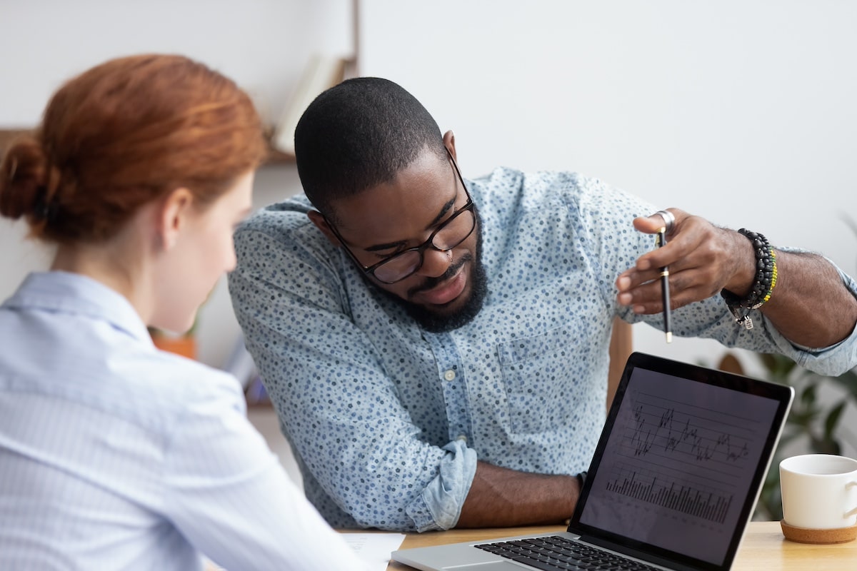 Diverse millennial colleagues working together analyzing diagram looking at computer screen. Black mentor helps female apprentice understand corporate program explaining interface showing on monitor -