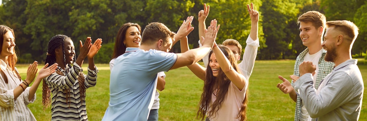 Eine Gruppe fröhlicher, vielfältiger Menschen feiert den Sieg in einem Outdoor-Teamspiel in einem grünen Sommerpark. Ein glücklicher junger Mann und eine junge Frau geben sich High Five, während ihre Freunde in die Hände klatschen und jubeln. -