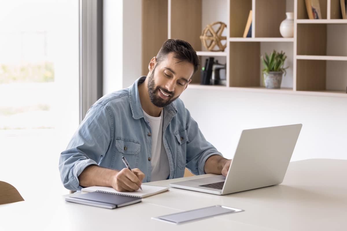 Positive Hispanic entrepreneur man using laptop for working in office, writing notes, smiling, talking on video call. Company manager, businessman watching professional learning webinar - workplace wellbeing