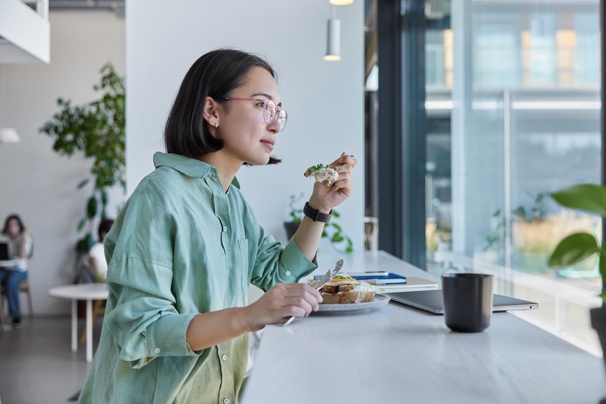 Junge, ethische Frau genießt leckeres Mittagessen, posiert in gemütlicher Cafeteria auf Fensterbank, umgeben von moderner Technologie für freiberufliche Tätigkeit, trägt Brille und legeres Hemd, blickt nachdenklich in die Zukunft