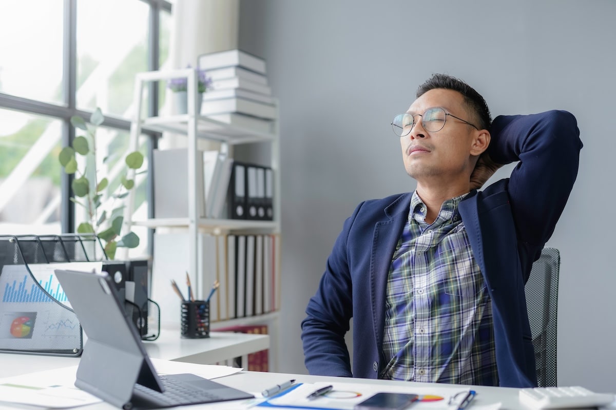 Asian businessman takes a moment to relax and stretch in his office, surrounded by paperwork and a laptop, finding relief and peace amidst the demands of his job - workplace wellbeing