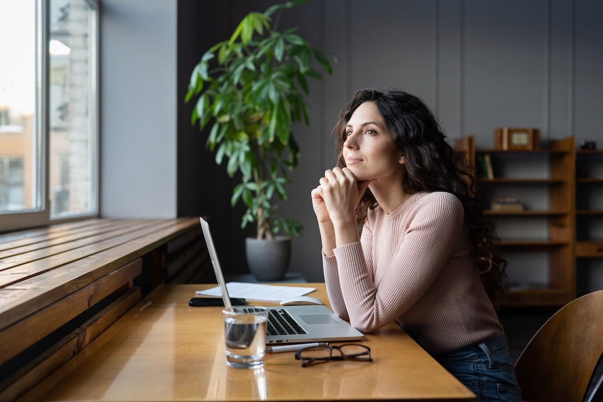 Young dreamy smiling female entrepreneur thinking about business goals and objectives, enjoying good window view while working in office, pleased businesswoman resting from computer work - motivation