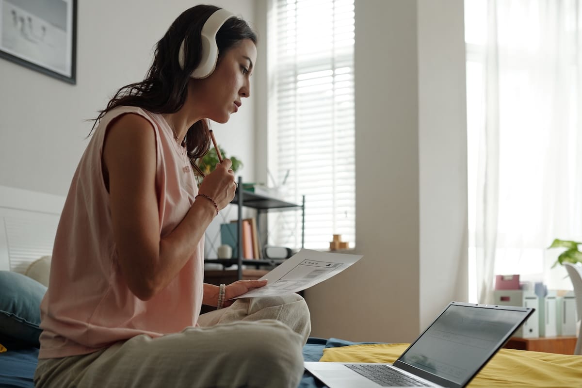 Woman sitting on floor by window, wearing headphones and holding document, working on laptop during daytime. Light from window streaming in softly - resilience