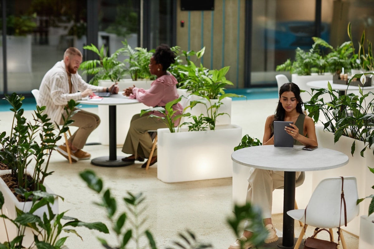 Wide angle shot of woman using tablet while sitting at table in cafe of office building. Business people working in cozy cafe featuring modern biophilic design - employee retention